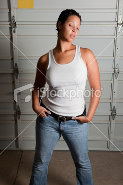 A photo of a medium skinned butch woman standing with her backdrop as what looks to be the inside of a white garage door. She is wearing a white tank top, low-waisted medium blue jeans, and a plain black leather belt. Her feet are not visible. she has very short dark hair, large breasts (though the tank top covers them well), and has her hands tucked into her front pockets with her thumbs hooked into the front two belt loops.