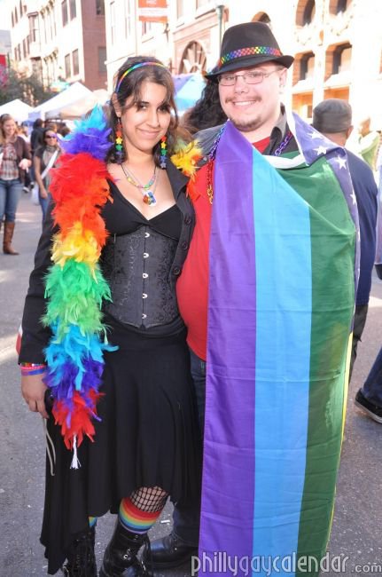  a black fedora with a rainbow band, a black collared shirt over a red shirt, a rainbow plastic chain, dark blue jeans, black boots, and a rainbow American flag that obscures most of his outfit