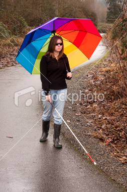A picture of a woman holding a large rainbow umbrella in her left and and a long white cane in her right hand. She is walking along a winding road and is wearing a long sleeve black shirt and light blue jeans that are tucked into dark grey rainboots. She is wearing sunglasses.
